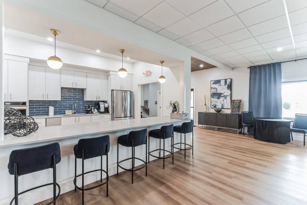 A spacious kitchen area with a long counter, blue backsplash tiles, modern white cabinets, and black bar stools in mesa az