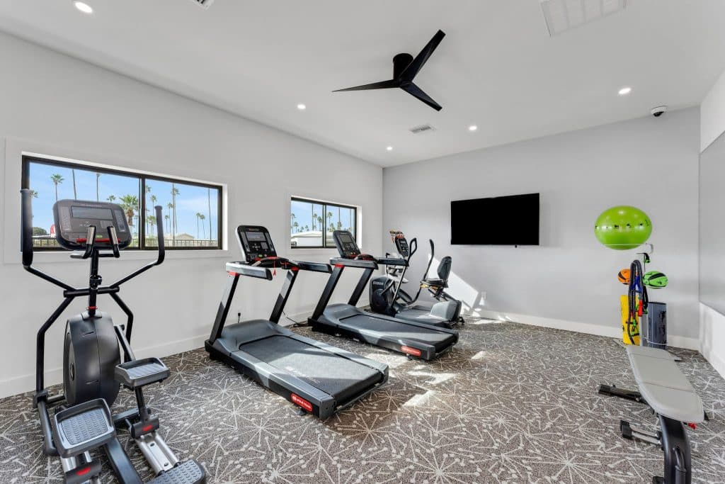 A fitness room with treadmills, an exercise bike, a bench, and a wall-mounted TV. Large windows let in natural light, showing palm trees outside in mesa az