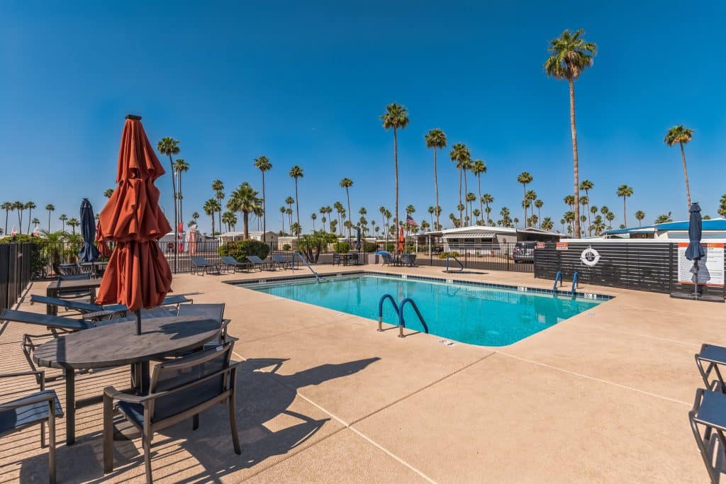A sparkling outdoor swimming pool surrounded by palm trees, lounge chairs, and red umbrellas under a bright blue sky, evoking a relaxing resort ambiance in mesa az